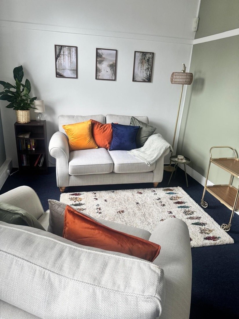 The counselling room at Within the Willow: a cream sofa with mustard, orange and navy cushions beneath three framed willow paintings